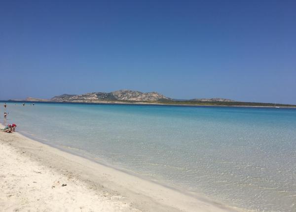 Spiaggia con sabbia bianca, mare cristallino e montagne sullo sfondo.