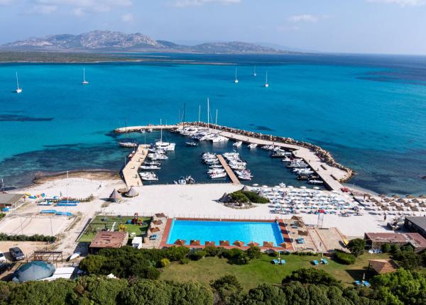 Porticciolo con barche su mare turchese, spiaggia e piscina, atmosfera di relax estivo.